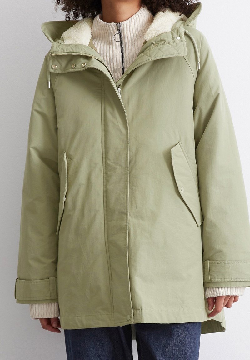 Veste imperméable vert clair avec capuche, col doublé en polaire, poches latérales et poignets ajustables. Le matériau a une texture lisse.
