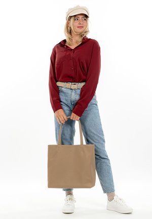 Frau mit beiger Mütze, burgunderroter Bluse, blauen Jeans, weißen Turnschuhen und großer beiger Tragetasche vor weißem Hintergrund.