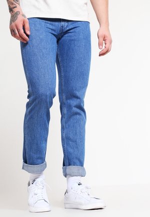 Mężczyzna noszący podwinięte niebieskie dżinsy, białe skarpetki i białe sneakersy Nike, stojący z widoczną tatuażem lewą ręką na białym tle.