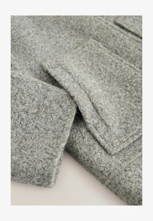 Pilkas vilnonis švarkas su minkšta, tekstūruota medžiaga. Turintis apykaklę ir kišenes, demonstruojantis šiek tiek pūkuotą paviršių ir pritaikytą dizainą.