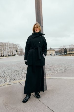 Svart ullrock med en oversized design, med en hög krage, två knappar och midjeband. Matchad med en lång svart kjol och stövlar.