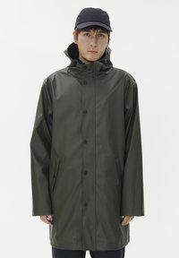 Grön vattentät regnjacka med huva, tryckknappar och sidofickor. Slät textur och avslappnad passform, kombinerad med en svart keps.