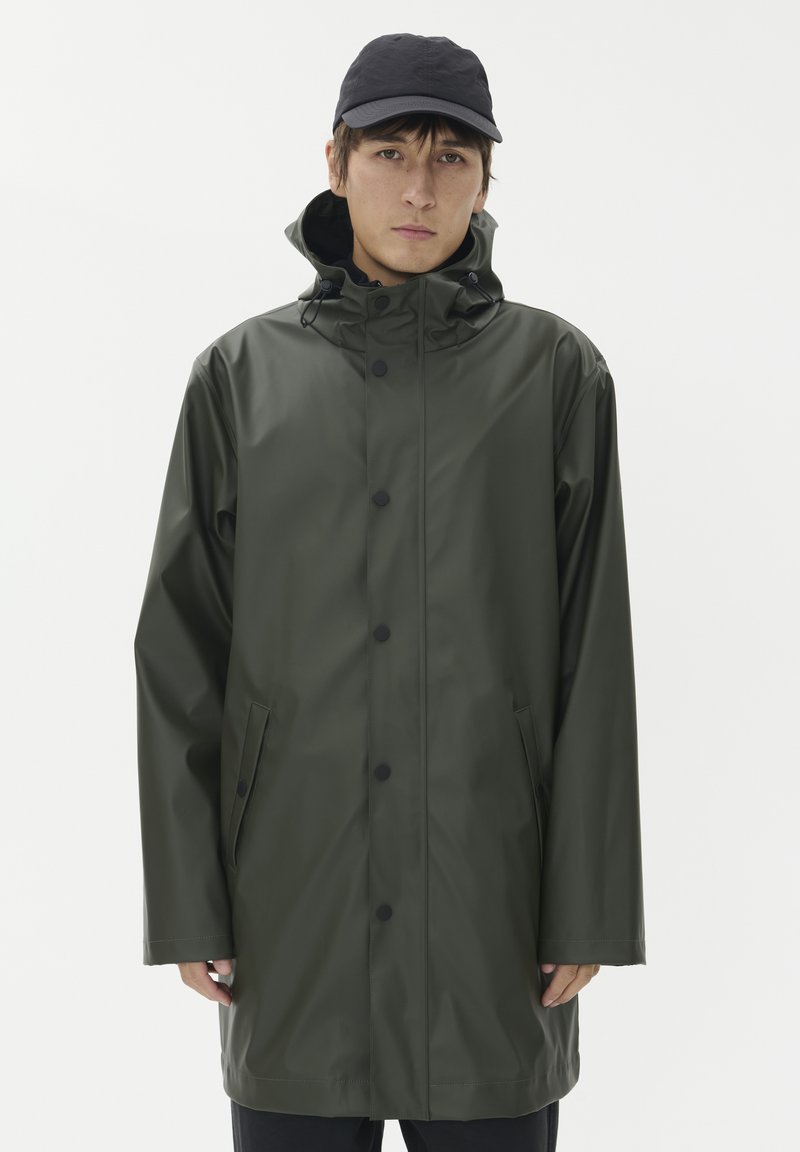 Grön vattentät regnjacka med huva, tryckknappar och sidofickor. Slät textur och avslappnad passform, kombinerad med en svart keps.