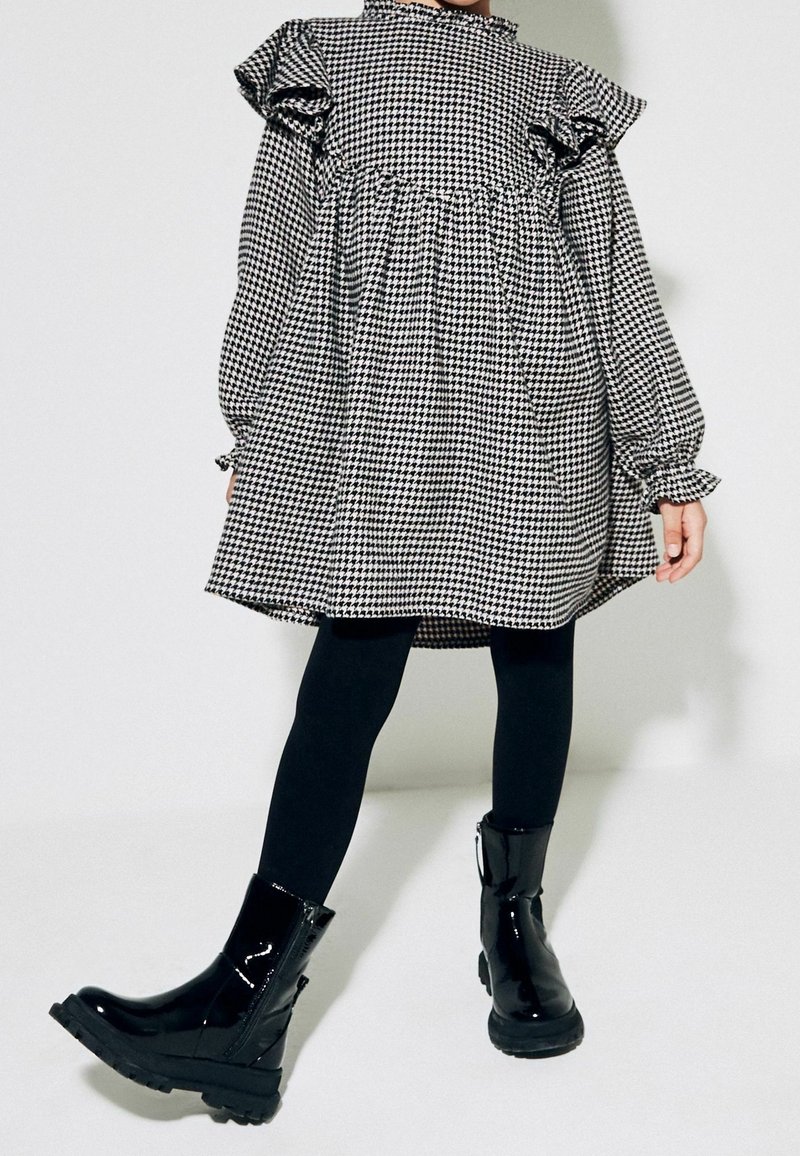 Enfant portant une robe pied-de-poule noire et blanche avec des volants, des leggings noirs et des bottines noires brillantes, debout sur un fond blanc.