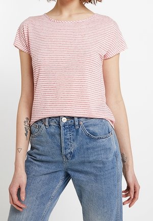 Frau trägt ein kurzärmeliges, rosa-weiß gestreiftes Oberteil, das in hoch taillierte blaue Jeans gesteckt ist, mit kleinen Tattoos auf beiden Unterarmen.