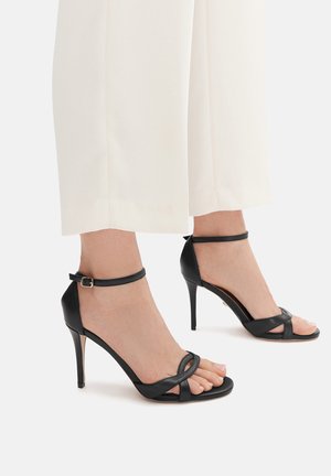 Sandali neri con tacco alto e punta aperta, caratterizzati da cinturini incrociati e una sottile fibbia alla caviglia. Finitura in pelle liscia.