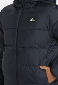 Námorná puffrová bunda so zipsom vpredu, s kapucňou, prešitými panelmi a malou nášivkou na hrudi. Mäkkučký, hladký nylonový materiál.