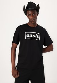 Juodas medvilninis marškinėliai su baltu stačiakampiu spaudiniu su žodžiu "oasis". Trumpomis rankovėmis ir apvaliu dekolte, dėvimi su juodomis kelnėmis.