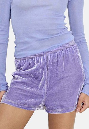 Vrouw draagt zachte paarse fluwelen shorts met een elastische taille en een bijpassende nauwsluitende top met lange mouwen, staand met ontspannen handen langs haar lichaam.