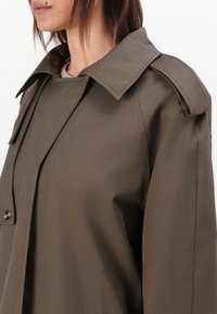 Palton khaki cu guler lat, prevăzut cu închidere cu nasturi și epoleți la umeri. Materialul neted, texturat oferă un aspect structurat.
