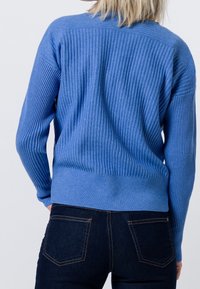 Modrý žebrovaný svetr s dlouhými rukávy, kulatým výstřihem a přiléhavým lemem. Nošený s tmavými džínovými kalhotami, ukazující texturovaný pletený vzor.