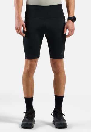 Jambes d'une personne portant un short de sport noir mi-cuisse, des chaussettes noires, des chaussures de course noires et une montre connectée au poignet gauche.