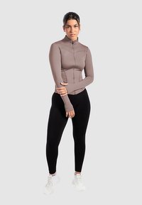 Strakke sportjas met ritssluiting in taupe, textuurbadstof met verhoogde naden, gecombineerd met zwarte legging en witte sneakers.