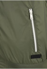 Tissu vert avec une poche zippée blanche verticale et une étiquette rectangulaire noire sur une veste ou un manteau.