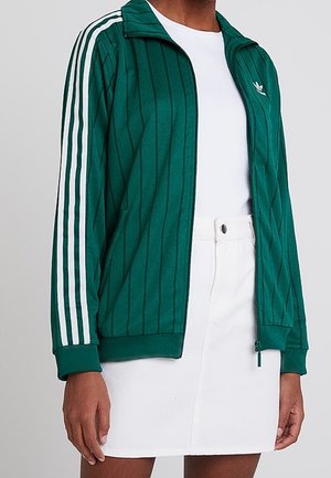 Personne portant une veste zippée verte avec des rayures blanches sur les manches, un haut blanc et une jupe blanche boutonnée, montrée du cou jusqu'au milieu des cuisses.