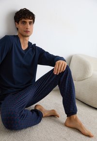 Marineblauwe longslavenshirt gecombineerd met gestreepte marineblauwe broek met rode accenten. Zacht materiaal, relaxte pasvorm en taps toelopende enkels.