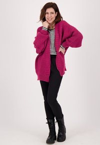Oversized gebreide cardigan in fuchsia, met een textured patroon, open voorkant en verlaagde schouders, gedragen over een zwart-wit gestreepte top en zwarte broek.