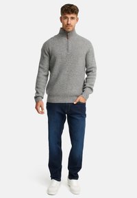Mand står op, iført en grå ribbet halv-zip sweater, mørkeblå jeans og hvide sneakers, med den ene hånd i lommen.