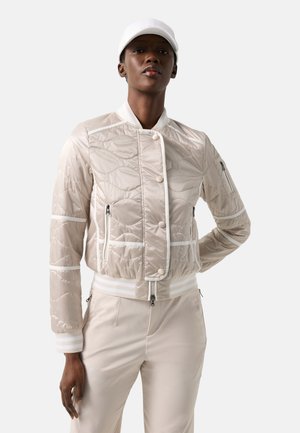 Kvinde iført en lysebeige quiltet jakke med hvid kant, matchende bukser og hvid baseballkasket mod en ensfarvet baggrund.