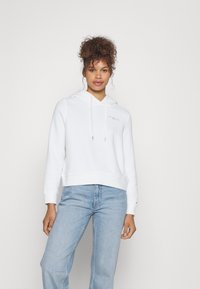 Felpa bianca con cappuccio regolabile e dettagli del logo frontale, abbinata a jeans azzurri chiari. Il tessuto appare morbido e strutturato.