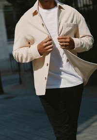 Beige Kordjacke mit geradem Schnitt, zwei Knöpfen und einer Brusttasche, getragen über einem weißen T-Shirt und schwarzen Hosen.