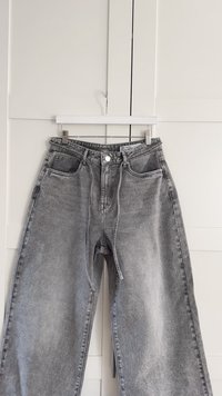 Γκρίζα τζιν παντελόνια με ψηλή μέση, φαρδιές γραμμές, μπροστινές τσέπες, θηλιές ζώνης και κλείσιμο με ασημένιο μεταλλικό κουμπί. Φθαρμένη υφή σε όλη την επιφάνεια.