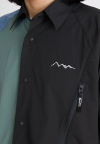 Black and green multi-panel button-up shirt, features a silver mountain logo, zip pocket, and smooth fabric texture with minimalistic design.