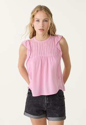 Pink sleeveless top with ruffled shoulders, textured front pleats, and a relaxed fit, paired with black denim shorts.