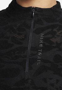 Černý sportovní top s půlzipem, vyrobený z texturované tkaniny, s výšivkou "NIKE TRAIL" na přední straně.