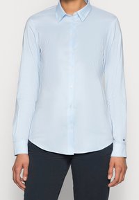Mujer vestida con una camisa de botón de manga larga azul claro y pantalones oscuros, de pie con las manos relajadas a los lados contra un fondo liso.