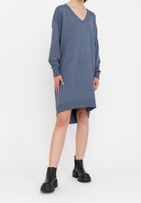 Vestito blu a V con maniche lunghe, polsini a coste e orlo asimmetrico. Realizzato in un tessuto morbido, indossato con stivali neri alla caviglia.