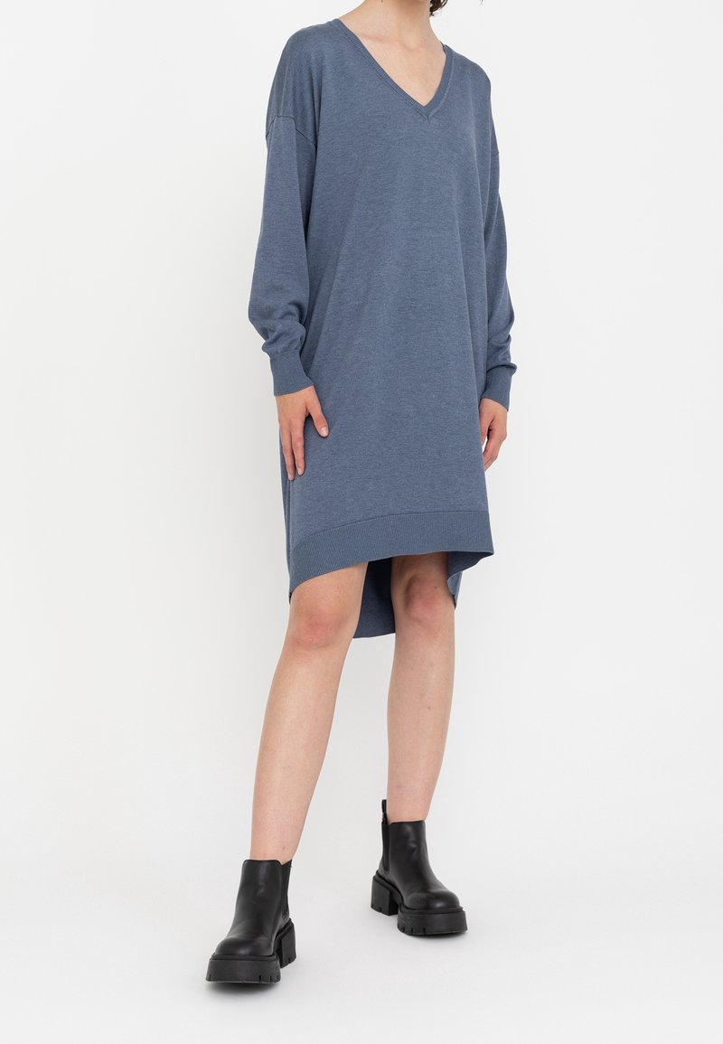 Vestito blu a V con maniche lunghe, polsini a coste e orlo asimmetrico. Realizzato in un tessuto morbido, indossato con stivali neri alla caviglia.