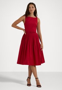 Rochie roșie fără mâneci, cu decolteu pătrat, bust ajustat, fustă plisată în formă de A și talie coordonată cu curea. Textură de țesătură netedă.