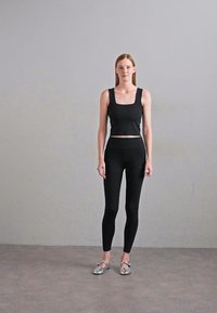 Top cropped negru, ajustat, cu bretele largi și colanți asortați. Modelul poartă pantofi slip-on argintii, stând pe un fundal neutru.