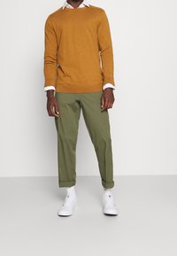 Maglione giallo senape in materiale lavorato a maglia, con scollo rotondo. Pantaloni di cotone verde, aderenti ai polsini, abbinati a sneakers bianche e calzini.