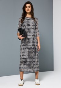 Mittellanges Kleid aus durchscheinendem grauem Stoff mit schwarzem abstraktem Muster. Es hat Dreiviertelärmel. Das Model hält eine schwarze Clutch und trägt weiße Sneaker.