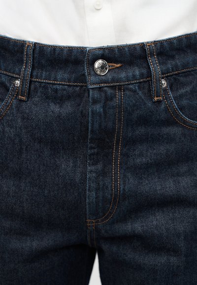 Close-up of dark blue denim jeans with orange stitching worn with a white button-up shirt tucked inside the waistband.