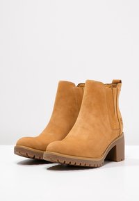 Vaaleanruskeat mokkanahka nilkkurit, joissa on pinottu korko, pyöreä kärki, sivuvetoketju ja teksturoitu kumipohja. Yksinkertainen ja puhdas muotoilu.