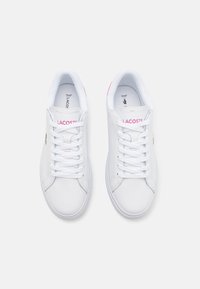 Bílé tenisky s hladkým koženým svrškem, kulatou špičkou a plochými bílými tkaničkami. Obsahují růžové akcenty a logo "LACOSTE" na boku.