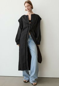 Woman wearing a long black buttoned coat over a black top, light blue wide-leg jeans, and black open-toe sandals standing against a white wall.