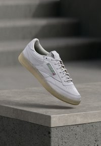 White leather sneaker with perforated toe, beige rubber sole, features green Reebok logo on the side, sleek design, minimal stitching details.