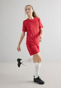 Jauna moteris futbolininkė raudonos uniformos, baltų kelio aukščio kojinių ir juodų sportinių batelių, pozuoja bėgimo metu priešais paprastą baltą foną.