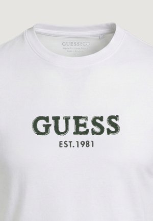 Balti marškinėliai su žaliu siuvinėtu „GUESS“ tekstu ir žemiau krūtinės užrašu „EST. 1981“.
