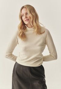 Beige kõrge kaelusega särk pehmest kangast, pikkade varrukatega ja liibuva disainiga, koos musta nahast seeliku ka.