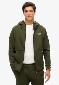 Felpa con zip verde oliva con accenti neri e logo, dotata di cappuccio con coulisse e due tasche frontali, abbinata a pantaloni jogger coordinati.