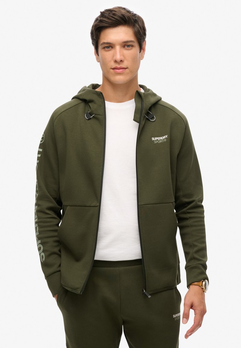 Felpa con zip verde oliva con accenti neri e logo, dotata di cappuccio con coulisse e due tasche frontali, abbinata a pantaloni jogger coordinati.