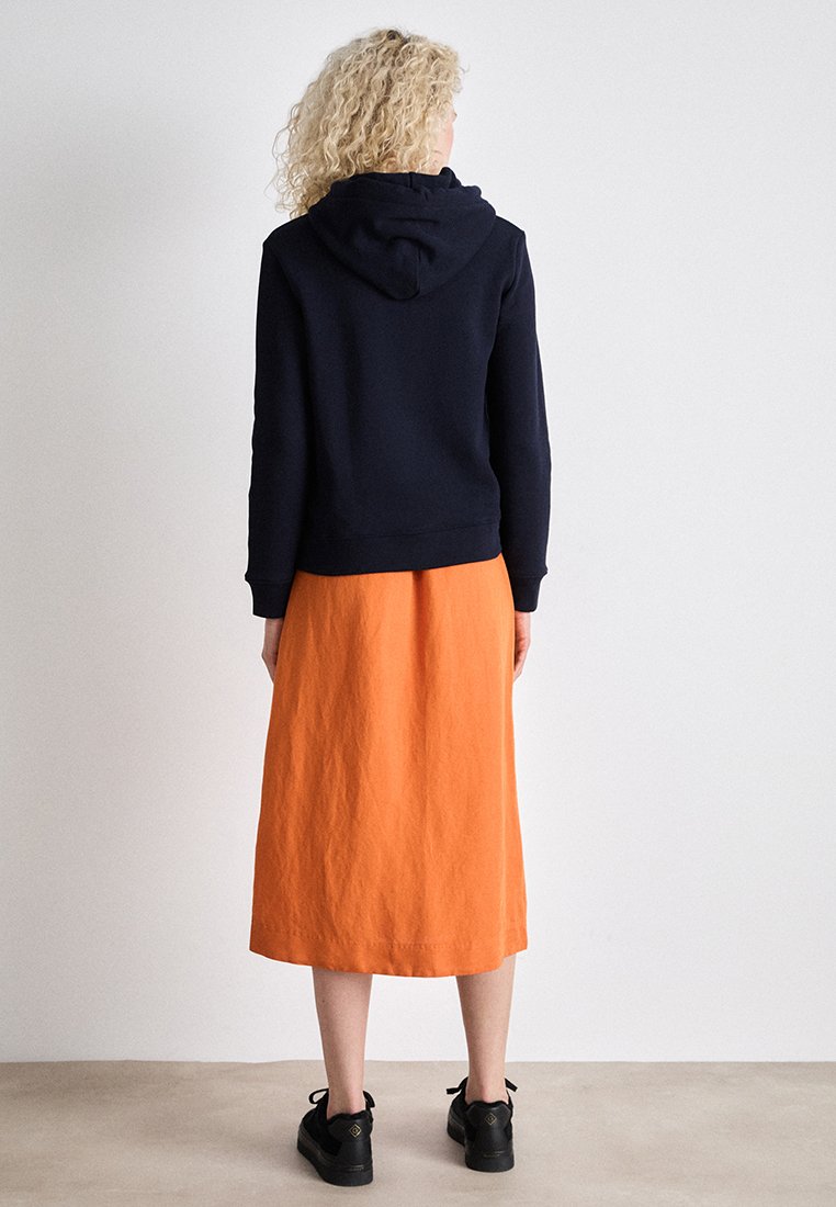 Frau mit lockigem blonden Haar, die einen schwarzen Hoodie, einen orangen Midirock und schwarze Sneaker trägt, steht vor einem einfarbigen hellen Hintergrund.