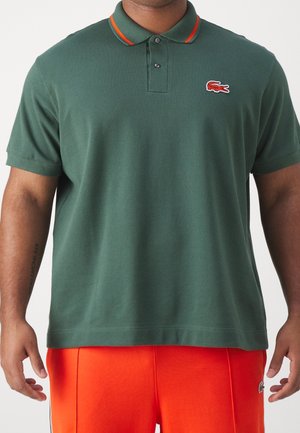 Homme portant un polo vert foncé avec des bordures orange et un pantalon orange vif, debout devant un fond blanc uni.