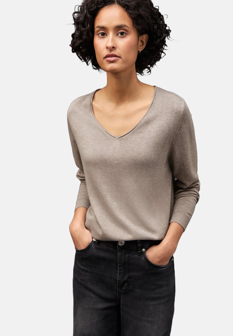 Donna con capelli neri ricci, che indossa un maglione beige a scollo a V e pantaloni neri, con le mani in tasca, in piedi contro uno sfondo chiaro e semplice.