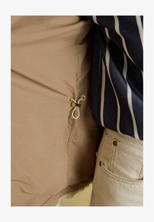 Beige Innenstoff mit einem goldenen Hardwarering und Toggle, unter einem marineblauen gestreiften Außenstoff geschichtet. Sichtbare weiße Textur am Saum.